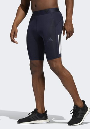 Mężczyzna ubrany w czarne spodenki kompresyjne Adidas z białymi paskami i czarne buty do biegania Adidas, stojący na jasnym, jednolitym tle.