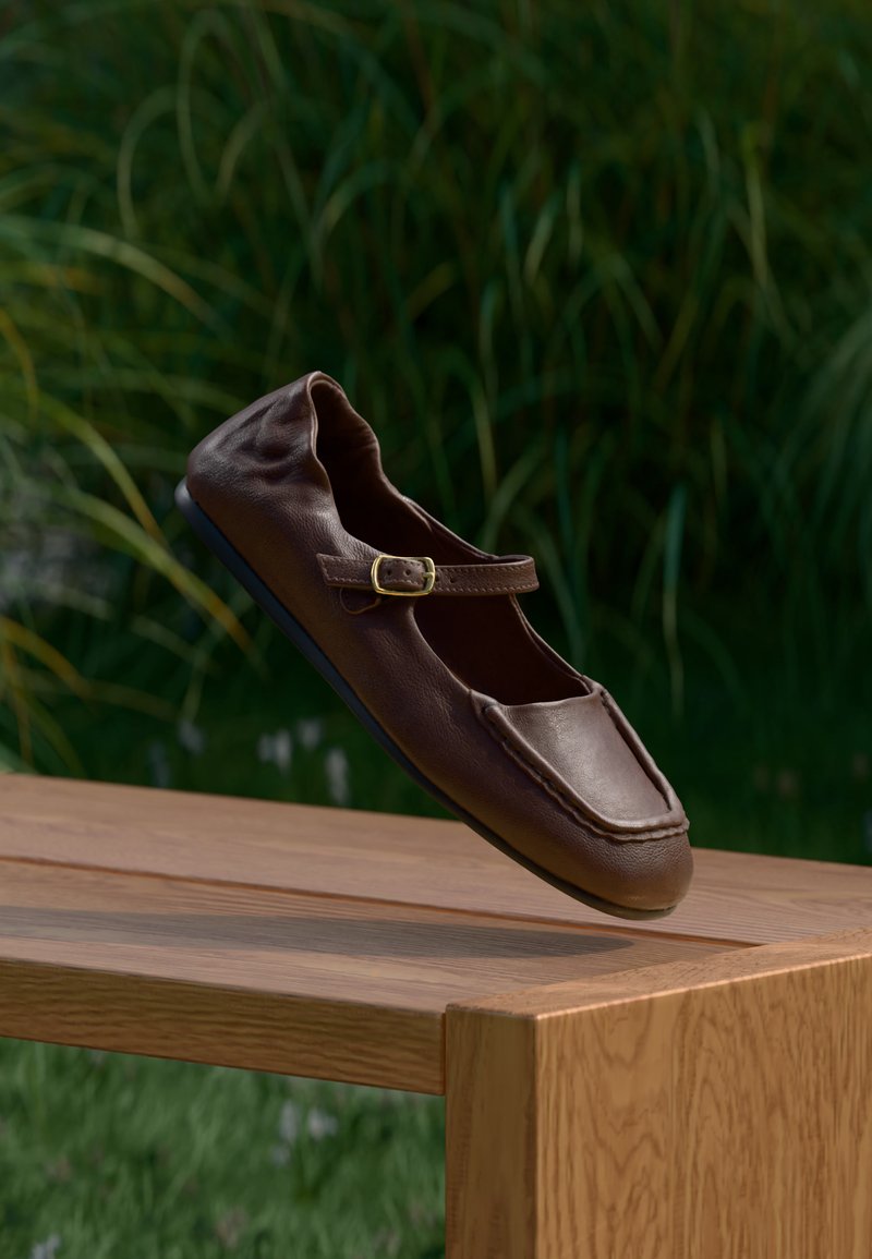 Chaussure plate Mary Jane en cuir marron avec bride à boucle, posée au-dessus d'une table en bois à l'extérieur avec un fond de feuillage vert.