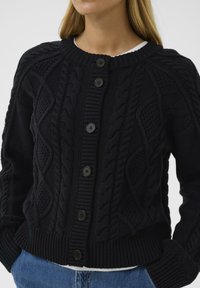 Fekete kötött kardigán, texturált kábelmintával, kerek nyakkivágással és hat gombbal elöl. Bordás mandzsetták és szegély.
