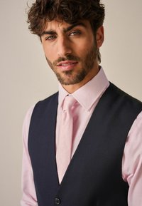 Gilet bleu marine sombre sur une chemise habillée rose pâle avec un motif subtil et une cravate assortie. Le tissu est texturé, ajoutant de la profondeur à l'ensemble.