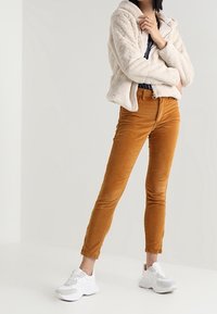 Beige Fleecejacke, marineblaues Oberteil, orangefarbene Kordhosen und weiße Turnschuhe. Die Hose ist schmal geschnitten; die Jacke hat einen Reißverschluss und eine strukturierte Stoffoberfläche.