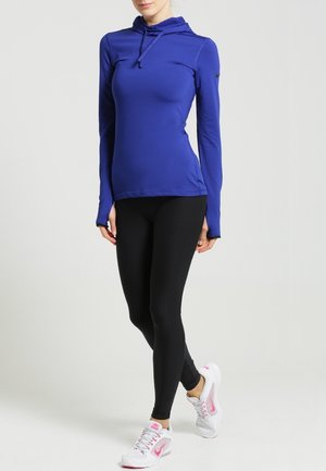 Vrouw die een nauwsluitende blauwe hoodie, zwarte legging en witte sneakers met roze accenten draagt, staand met één been gekruist aan de voorkant.