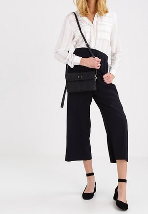 Sort håndtaske med vævet tekstur, justerbar strop og metal hardware. Modellen bærer en hvid stribet skjorte med knapper og sorte cropped bukser.