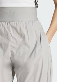 Svetlo šedé športové nohavice s elastickým pásom, na ktorom sa nachádza jemne reliéfne logo. Materiál vyzerá hladký a trochu pokrčený.