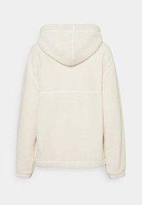 Krämfärgad fleecejacka med huva, med en mjuk struktur, sidofickor och synliga sömmar på baksidan.