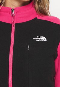 Svart fleecejacka med livfulla rosa ärmar. Har en dragkedja fram, hög krage och vit broderad logotyp på bröstet.