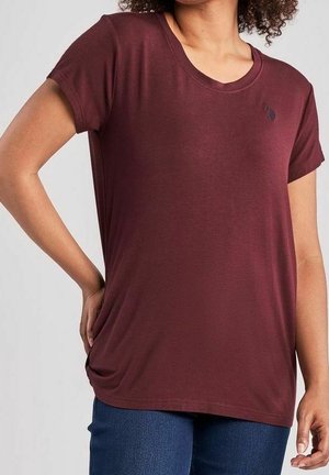 Kobieta ubrana w bordowy t-shirt z krótkim rękawem i małym logo na lewej piersi, w niebieskich dżinsach, stojąca z jedną ręką na biodrze.