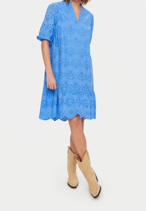 Kobieta w niebieskiej sukience z ażurowym wzorem i bufkami na rękawach oraz beżowych kowbojskich butach, stojąca ze skrzyżowanymi nogami na białym tle.