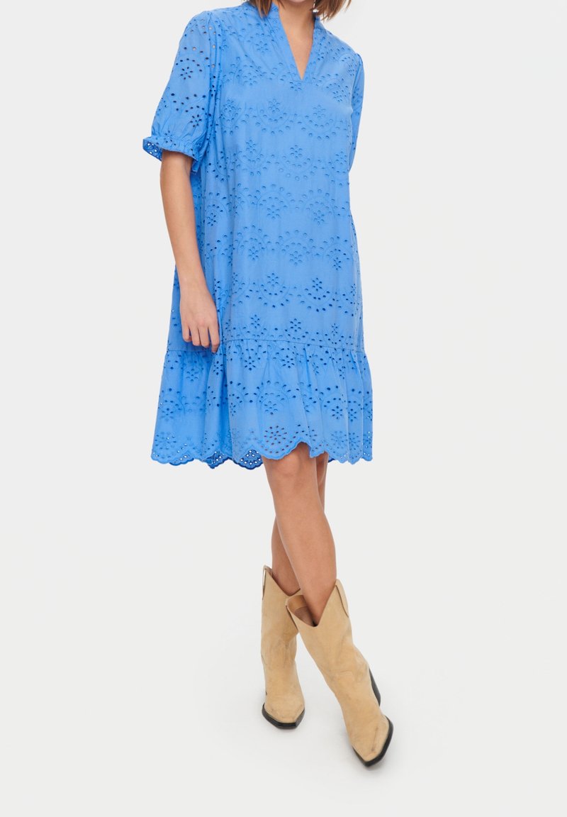 Femme portant une robe bleue en œillets avec des manches bouffantes et des bottes de cowboy beiges, debout les jambes croisées devant un fond blanc.