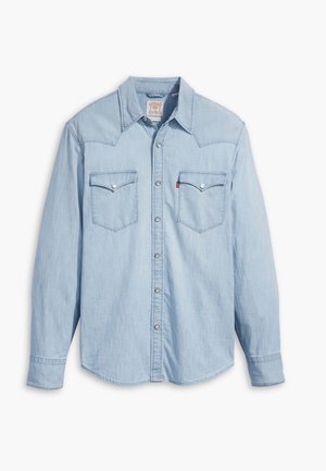 Chemise en denim bleu clair à manches longues avec boutons-pression et deux poches poitrine à rabats, posée à plat sur un fond blanc.