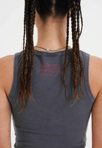 Débardeur gris sans manches avec un tissu texturé et un logo imprimé en rouge dans le dos, complété par des cheveux tressés et un collier.