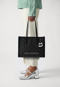 Juodas prekybinis krepšys su logotipu ir raštuota danga, su dviem rankenomis ir katės pakabuku. Derinama su sidabriniais aukštakulniais bateliais ir grietinėlės spalvos kelnėmis.