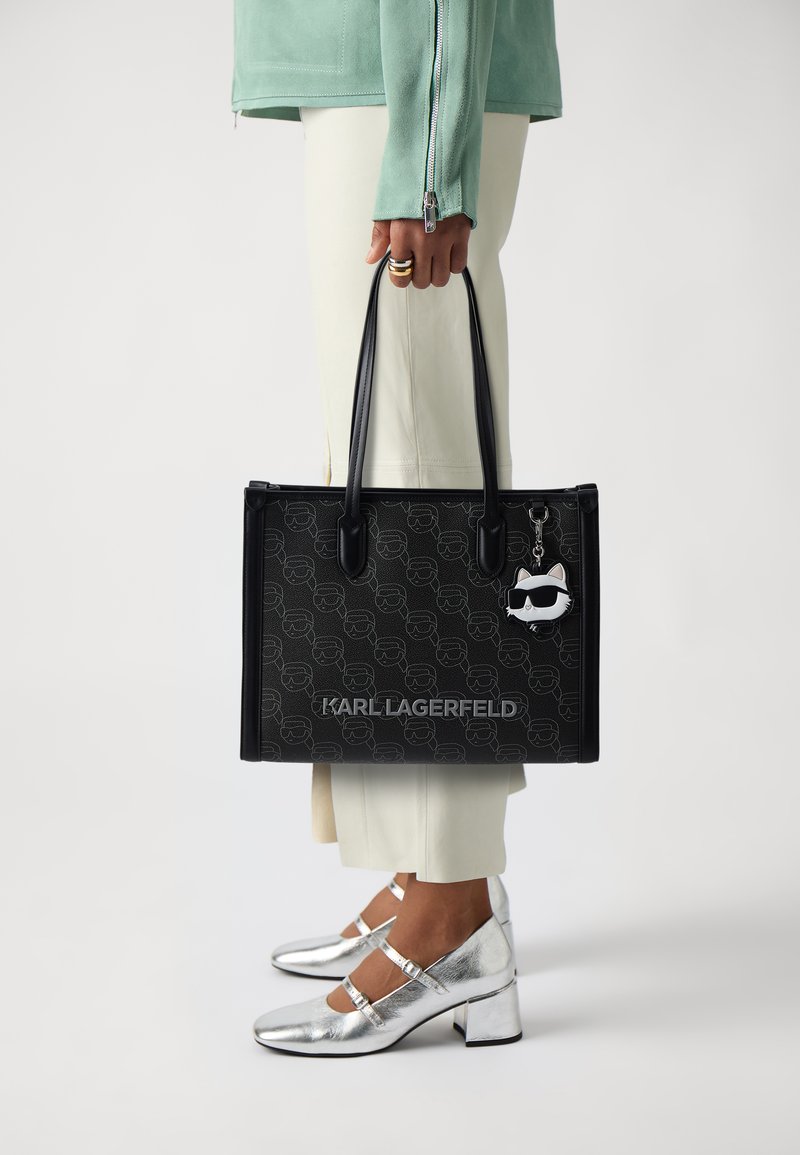 Juodas prekybinis krepšys su logotipu ir raštuota danga, su dviem rankenomis ir katės pakabuku. Derinama su sidabriniais aukštakulniais bateliais ir grietinėlės spalvos kelnėmis.