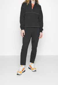 Sudadera negra con cremallera y costuras contrastantes en gris, combinada con pantalones deportivos negros con piernas estrechas y bolsillos laterales. Zapatillas llamativas.
