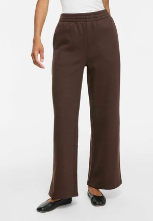 Femme portant un pantalon large marron avec une taille élastique et des chaussures plates noires, debout devant un fond blanc.