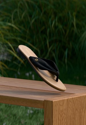 Black fabric thong sandal with beige sole balanced on edge of wooden bench outdoors with green plants in the background.