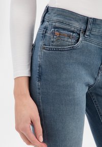 Jean en denim bleu à taille haute, avec une petite poche zippée et une finition texturée. Des poches latérales pour les mains sont visibles.