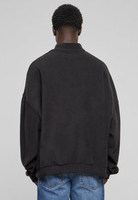Svart oversized sweatshirt med høy hals, nedfalne skuldre og ribbestrikkede mansjetter. Myk tekstur og minimalistisk design. Brukt sammen med blå jeans.