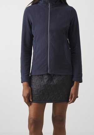 Femme portant une veste polaire bleu marine zippée et une mini-jupe matelassée noire, debout devant un fond clair uni.