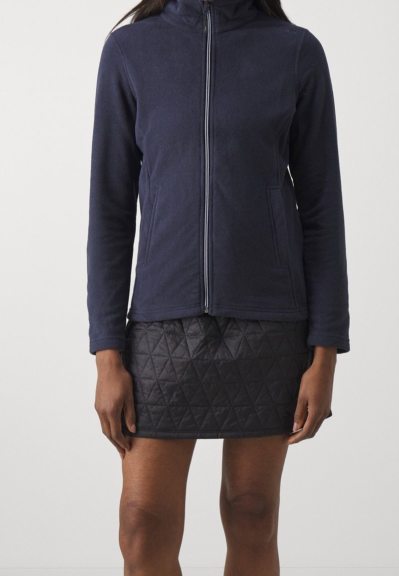 Femme portant une veste polaire bleu marine zippée et une mini-jupe matelassée noire, debout devant un fond clair uni.
