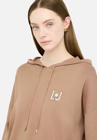 Brūna kapuce ar priekšējo kabatu, kurai ir kristāla rotāts "LJ" logo. Mīksts audums un auklas ar metalizētām galiem.