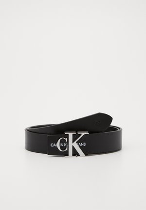 Ceinture en cuir noir avec boucle argentée "CK" et inscription "Calvin Klein Jeans" en relief sur la lanière, enroulée sur un fond blanc.