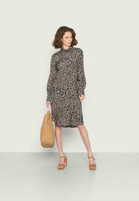 Vestido de manga longa, até ao joelho, com um padrão animal em preto e bege, apresentando um colarinho alto e um ajuste descontraído, combinado com uma bolsa bege e saltos altos.