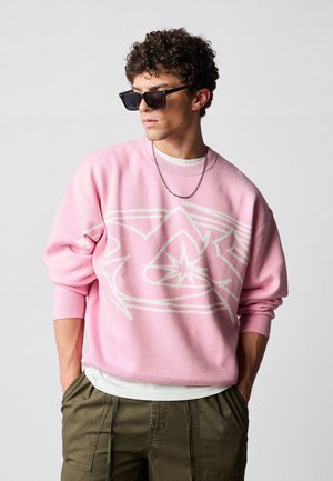 Jeune homme aux cheveux bouclés portant des lunettes de soleil noires, un pull rose avec un motif blanc, une chemise blanche en dessous et un pantalon vert olive, les mains dans les poches.