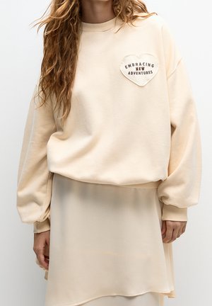 Kvinde iført en cremefarvet sweatshirt med et hjerteformet mærke med teksten "Embracing New Adventures" og et matchende cremefarvet nederdel.
