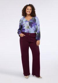 Donna con capelli ricci che indossa una blusa floreale blu e viola infilata in pantaloni a vita alta color bordeaux e tacchi a punta beige, sorridente.