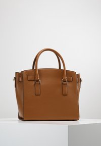 Borsa a tracolla in pelle marrone con doppio manico e minimo hardware in oro, appoggiata su un piedistallo bianco contro uno sfondo chiaro e semplice.