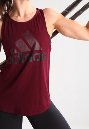 Personne portant un débardeur Adidas bordeaux et un legging noir, tenant une barre derrière les épaules dans une posture d'entraînement.