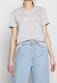 Mujer usando una camiseta gris de Calvin Klein metida dentro de unos pantalones vaqueros de talle alto de color azul claro, vista desde el cuello hasta las caderas.