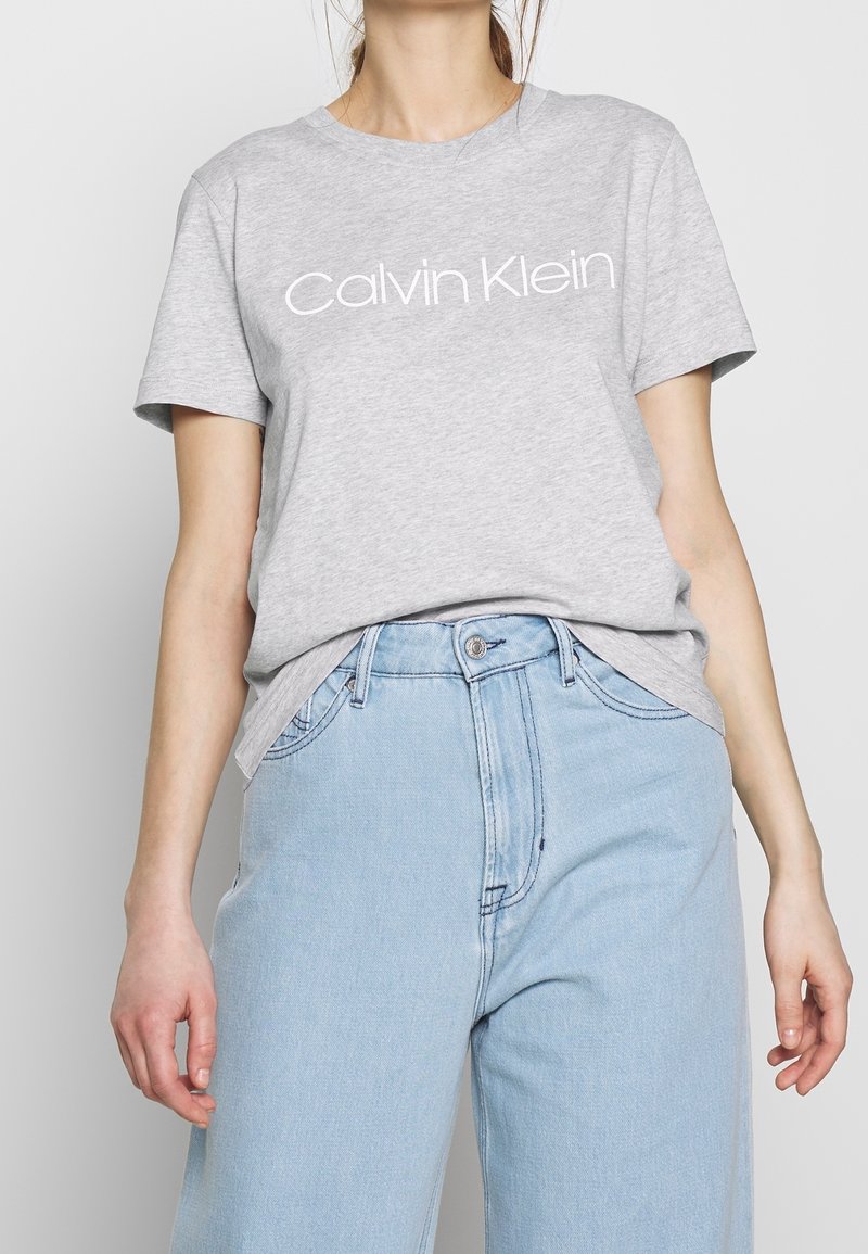 Mujer usando una camiseta gris de Calvin Klein metida dentro de unos pantalones vaqueros de talle alto de color azul claro, vista desde el cuello hasta las caderas.