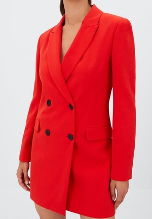 Femme portant une robe blazer rouge vif à double boutonnage avec des boutons noirs et des poches à rabat, debout devant un fond clair uni.