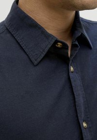Camisa de lino azul marino con cuello extendido, que presenta una textura suave, costuras visibles y un botón de latón en el cuello.