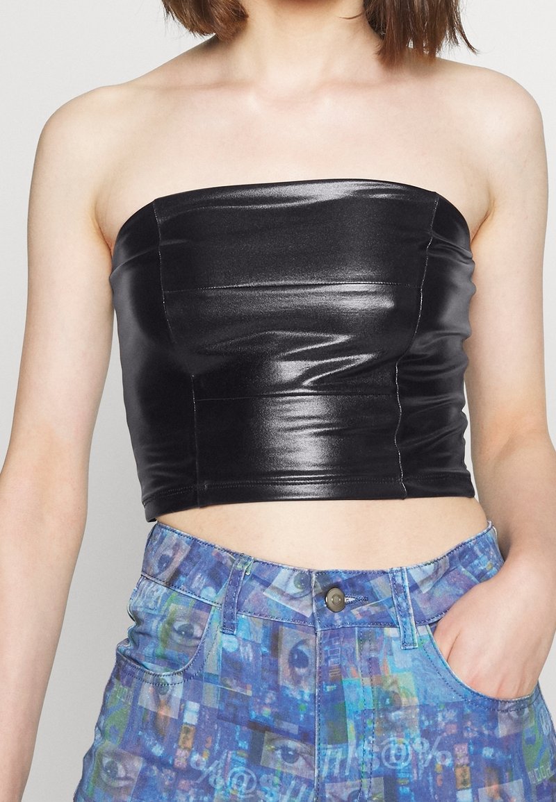 Femme portant un crop top noir sans bretelles brillant et un jean bleu à motifs taille haute, une main dans la poche.