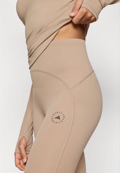 Μπεζ, εφαρμοστά κολάν με ψηλή μέση, με επίπεδες ραφές και κυκλικό λογότυπο στο αριστερό μηρό. Ομαλή υφή.