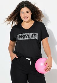 Zwarte V-hals t-shirt met witte tekst "POWER." Model houdt een roze bal vast en draagt losse zwarte broek met een trekkoord.