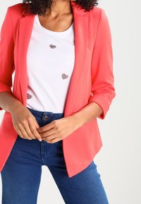 Vrouw draagt een koraalkleurig colbert over een wit t-shirt met drie glinsterende harten, gecombineerd met een blauwe spijkerbroek, handen samengevouwen bij de taille.