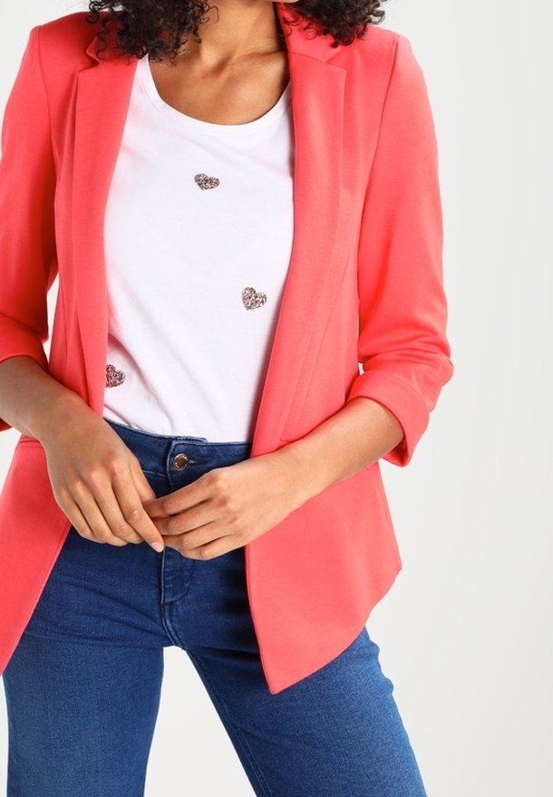 Vrouw draagt een koraalkleurig colbert over een wit t-shirt met drie glinsterende harten, gecombineerd met een blauwe spijkerbroek, handen samengevouwen bij de taille.