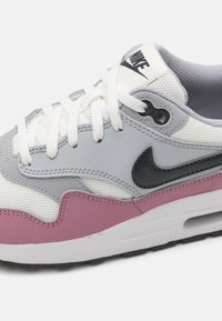 Nike Air Max 1 omab pehmeast võrkkangast ja nahast ülaosa hallis, valges ja roosas, mustade aktsentide ning valgete paeltega. Külgedel on tekstuuriga detailid.