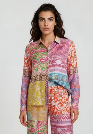 Frau trägt eine bunte Patchwork-Bluse und passende Hose mit Blumen- und Paisley-Mustern.
