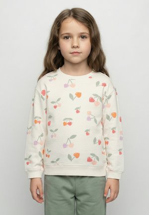 Sweat-shirt crème clair avec un imprimé de cerises en rose, rouge et vert. Présente un col rond et des poignets côtelés. Porté avec un pantalon vert.