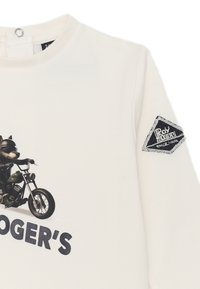 Felpa bianca con grafica di un lupo cartone animato su una motocicletta. Include un'etichetta nera testurizzata sulla manica con "Poy Rogers".