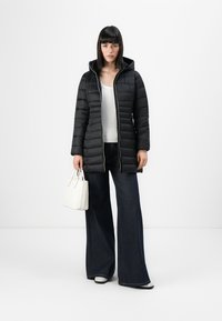 Zwarte pufferjack met een capuchon, horizontale gestikte textuur, voorzijde met ritssluiting. Gecombineerd met high-waisted donkerblauwe flare jeans en een witte handtas.