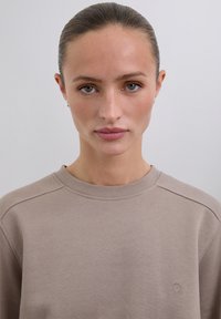 Junge Frau mit zurückgebundenem Haar, die einen beigen Sweatshirt mit einem kleinen Totenkopf- und gekreuzten Knochen-Logo auf der Brust trägt, neutraler Hintergrund.