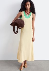 Femme aux longs cheveux bouclés portant une robe beige sans manches par-dessus un haut vert, tenant un grand sac à main marron en tricot, debout sur un sol gris.