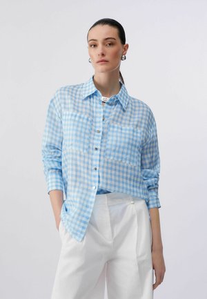 Žena s tmavými vlasmi stiahnutými do copu, oblečená v svetlomodrej károvanej košeli na gombíky s náprsnými vreckami a bielych vysokých nohaviciach, s rukami vo vreckách.