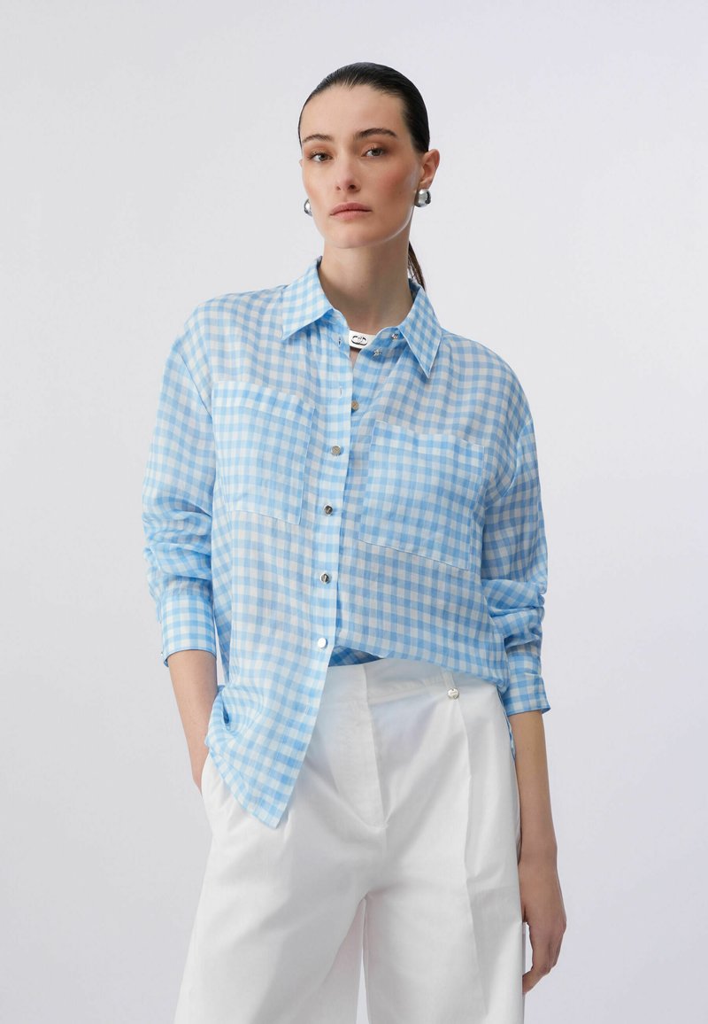 Femme aux cheveux foncés attachés en queue de cheval, portant une chemise à carreaux bleu clair boutonnée avec poches poitrine et un pantalon blanc taille haute, les mains dans les poches.